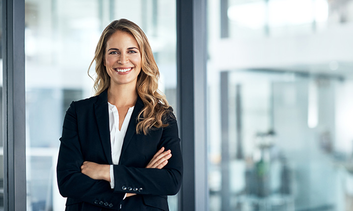 Woman wearing a blazer smiling and standing in front of a window in an office