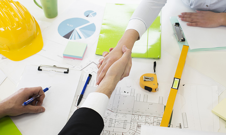 Two people shaking hands over a table with charts and planning materials on it
